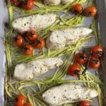 Baked chicken pieces seasoned with pepper are arranged on a bed of thinly sliced asparagus in a baking dish. Clusters of vine cherry tomatoes are placed among the chicken and asparagus.