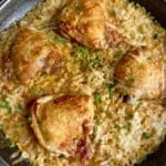 A skillet filled with golden-brown chicken thighs resting on a bed of creamy, baked rice speckled with fresh chopped herbs. The dish has a crispy and appetizing appearance, with a wooden spoon partially visible in the background.