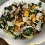 A salad in a white bowl featuring kale, roasted butternut squash, fennel slices, dates, seeds, and shaved parmesan on top. The dish is placed on a wooden cutting board.