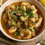 A bowl of shrimp in a rich, red sauce garnished with chopped parsley. Surrounding the bowl are slices of crusty bread and a lemon wedge. Two gold-colored utensils are placed neatly nearby.