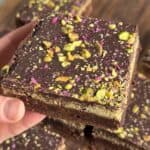 A close-up of a hand holding a layered dessert square. It features a chocolate top sprinkled with pistachios and pink rose petals, a cream filling, and a solid base. More identical squares are visible in the background on a wooden surface.
