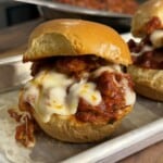 A meatball slider with melted cheese and tomato sauce on a shiny bun, served on a metal tray. Another slider and a pan with more filling are slightly blurred in the background.