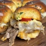 Close-up of a slider sandwich with melted cheese, sliced beef, and a slice of tomato on a wooden board. The sandwich bun is glossy and sprinkled with herbs. Other similar sandwiches are visible in the background.
