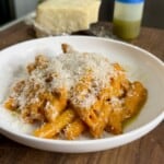 A white plate filled with pasta covered in a creamy orange sauce, topped with grated cheese. In the background, there is a block of cheese and a bottle labeled Basil Oil, placed on a wooden surface.