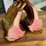 A perfectly cooked rack of lamb cut into two sections, displaying a pink, juicy interior and seasoned crust, resting on a wooden cutting board in a kitchen setting.