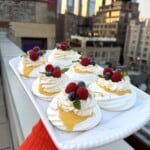 A person holds a rectangular platter of mini pavlovas topped with whipped cream, lemon curd, raspberries, blueberries, and mint on a rooftop, with a city skyline in the background at sunset.