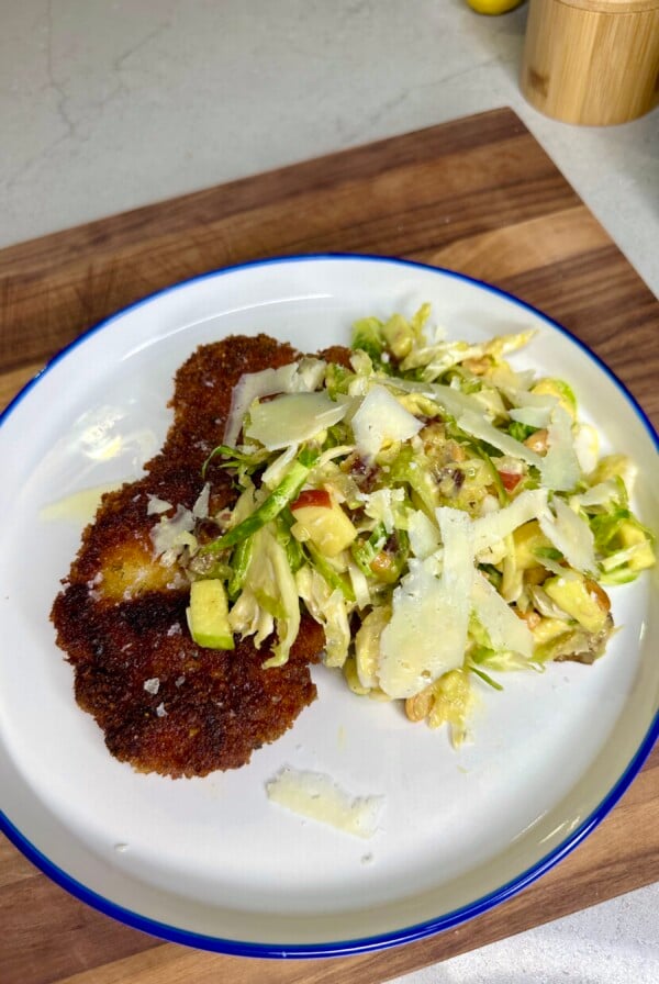A white plate with a blue rim holds two pieces of breaded, fried meat alongside a fresh cabbage salad topped with shaved cheese. The plate rests on a wooden cutting board.