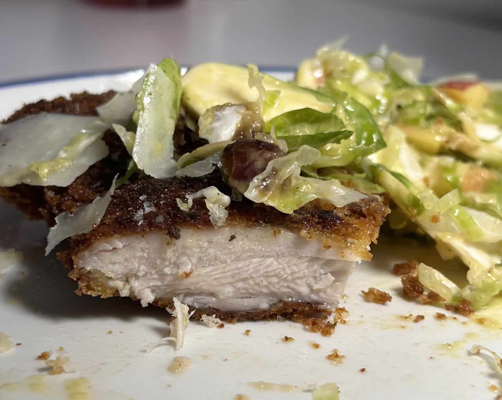 Close-up of a plate with a piece of breaded, cooked chicken cut in half, showing the white interior, next to a creamy salad with lettuce, avocado, and bacon.