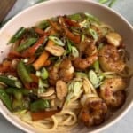A bowl of noodles topped with sautéed shrimp, sliced carrots, snap peas, red bell peppers, green onions, and crispy garlic chips on a light-colored surface.