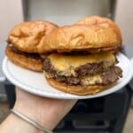 A hand holds a white plate with two cheeseburgers, featuring melted cheese and juicy beef patties in glossy buns. The background shows a blurred grill.