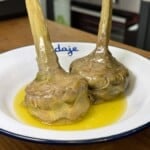 Two artichokes cooked and served upright in a shallow dish with olive oil, placed on a white plate with a blue rim on a wooden countertop in a kitchen setting.