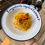 A hand holds a plate of spaghetti with clams, topped with grated cheese and herbs. The plate rim reads “L’ingrediente segreto è l’amore” with a small red heart. A street is visible below in the background.