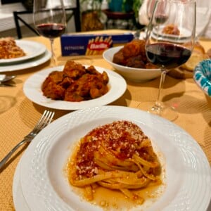 A plate of fettuccine pasta with tomato sauce and grated cheese sits on a table set for dinner, alongside red wine, a bowl of meatballs, and a box of Barilla fettuccine.