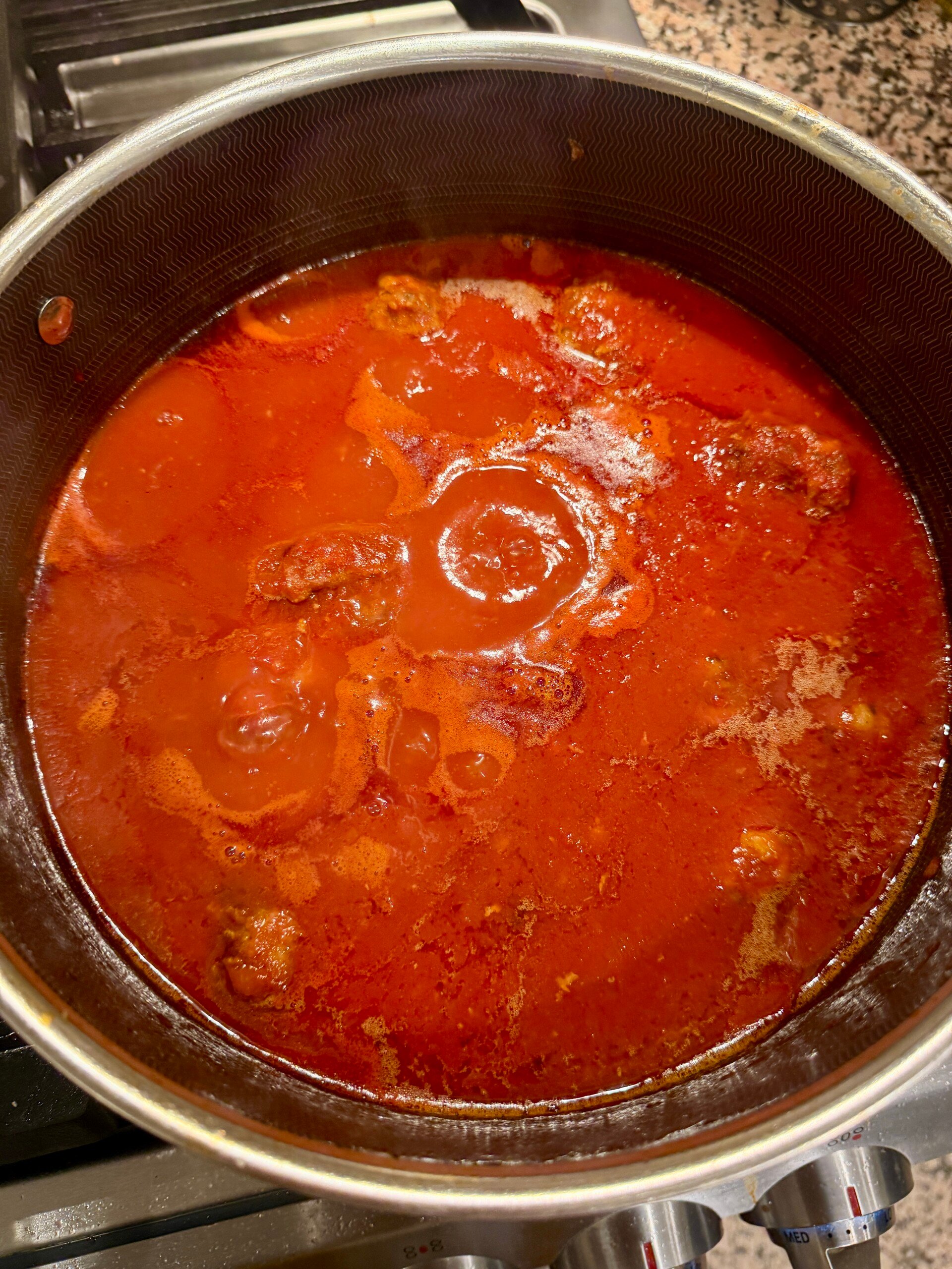 A pot of red tomato-based sauce with meat simmering on a stovetop. The sauce has visible oil on top and steam is rising as it cooks.