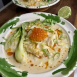 Two bowls of creamy soup with bok choy, fish, orange salmon roe, sliced green onions, and a drizzle of chili oil, served on green and white ceramic plates with a lime wedge on the side.