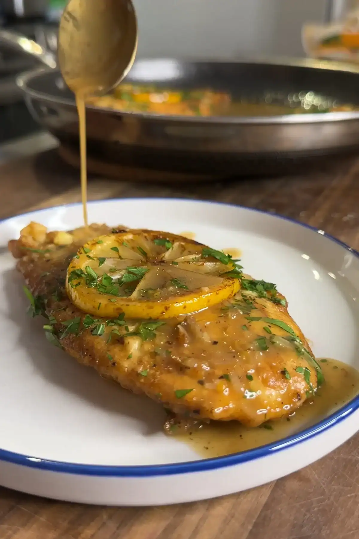 A piece of cooked chicken breast topped with a lemon slice and herbs is served on a white plate. Brown sauce is being spooned over the chicken. A skillet with more food is blurred in the background.