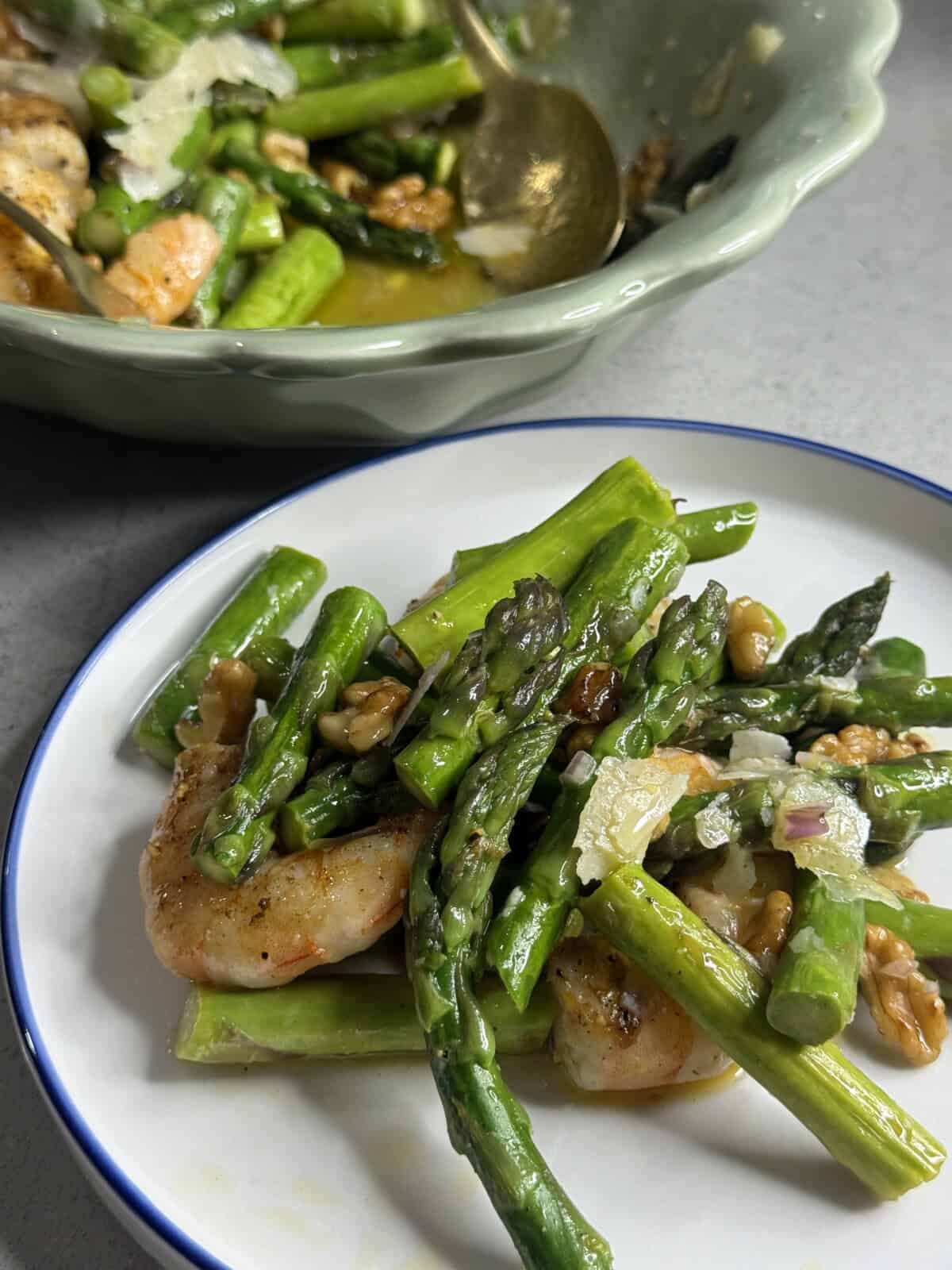 A plate of sautéed shrimp, asparagus, walnuts, and shaved parmesan, with a serving bowl containing more of the dish in the background. The meal is garnished with sauce and herbs.