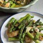 A plate of sautéed shrimp, asparagus, walnuts, and shaved parmesan, with a serving bowl containing more of the dish in the background. The meal is garnished with sauce and herbs.