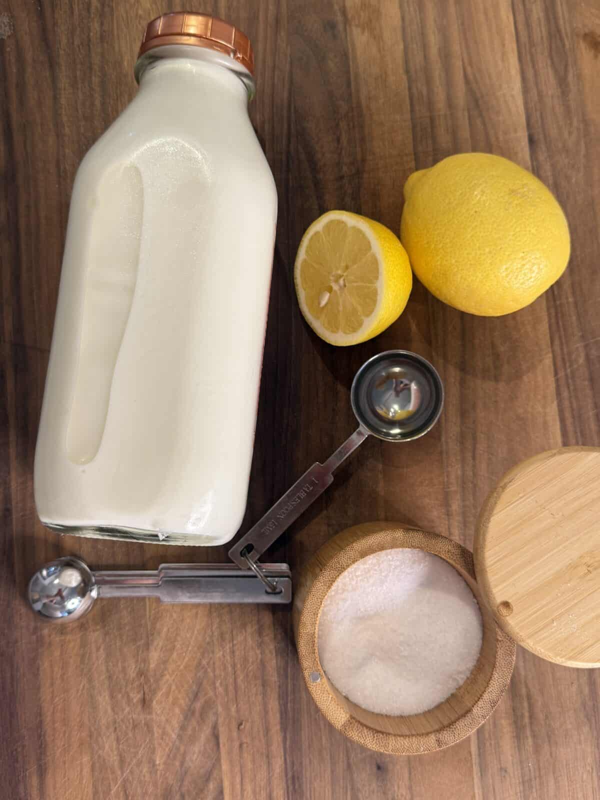 A bottle of milk, a small wooden bowl of salt, two lemons (one cut in half), and a metal measuring spoon are arranged on a wooden surface.