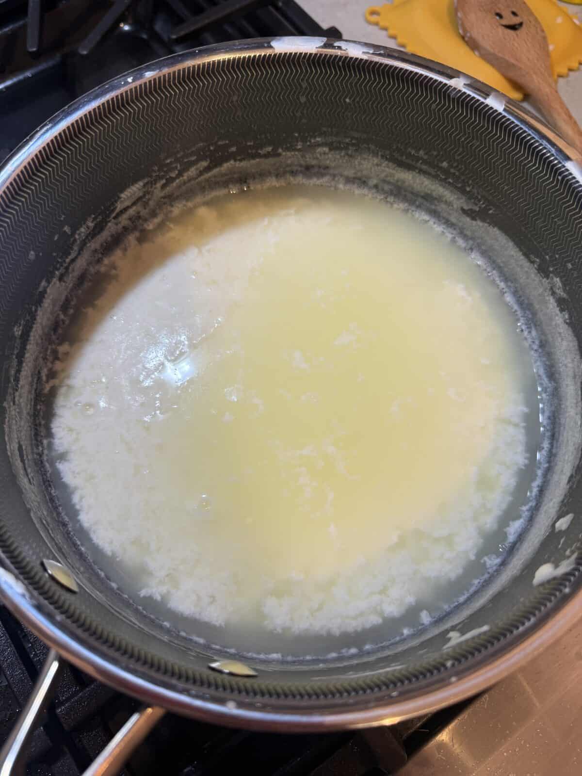 A saucepan on a stove contains curdled milk, with white curds separating from the yellowish whey during the process of making homemade cheese or ricotta. A wooden spoon and silicone trivet are nearby.