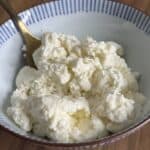 A bowl of ricotta cheese with a gold spoon on a wooden surface. The ricotta has a soft, crumbly texture and is white in color. The bowl has blue stripes along the rim.