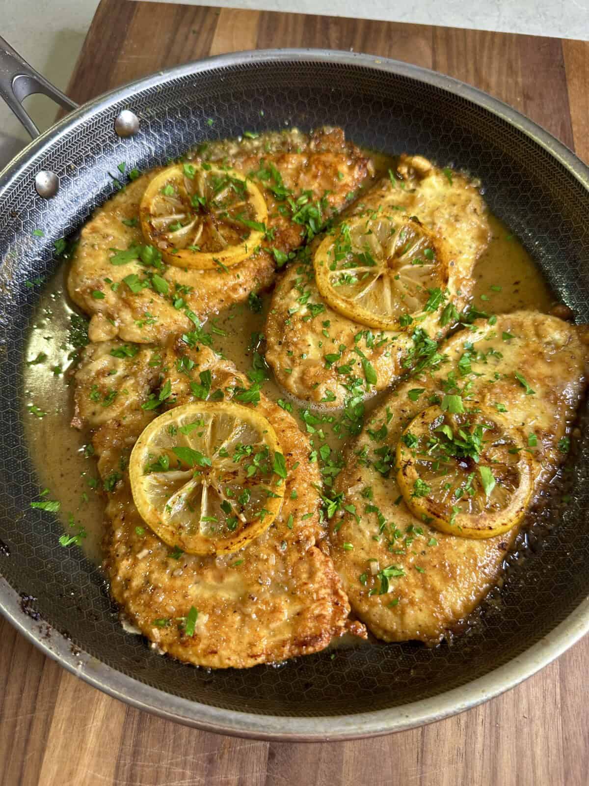Chicken cutlets cooked in a skillet, topped with lemon slices and garnished with fresh chopped parsley, sitting in a golden sauce on a wooden surface.