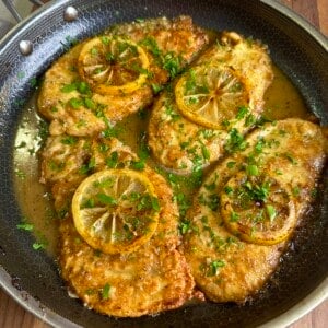 Chicken cutlets cooked in a skillet, topped with lemon slices and garnished with fresh chopped parsley, sitting in a golden sauce on a wooden surface.