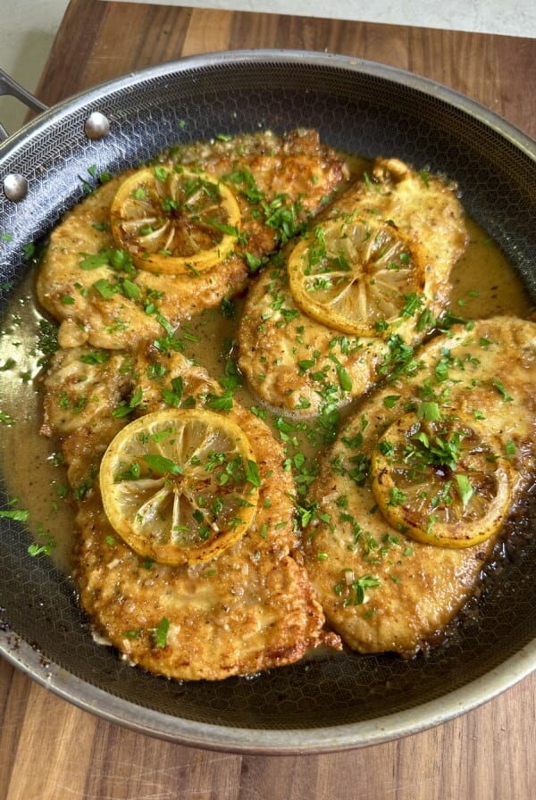 Chicken cutlets cooked in a skillet, topped with lemon slices and garnished with fresh chopped parsley, sitting in a golden sauce on a wooden surface.