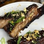 Two glazed, sliced barbecue ribs topped with chopped peanuts and green onions are served on a white plate. The ribs appear tender and caramelized, with a shiny, sticky sauce.