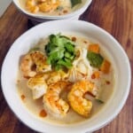 Two white bowls filled with shrimp soup, rice noodles, zucchini, carrots, fresh cilantro, and a drizzle of chili oil, served on a wooden surface.