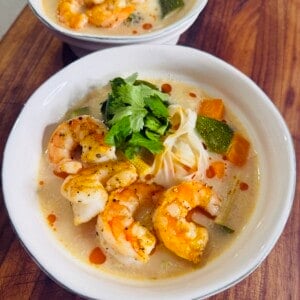 Two white bowls filled with shrimp soup, rice noodles, zucchini, carrots, fresh cilantro, and a drizzle of chili oil, served on a wooden surface.