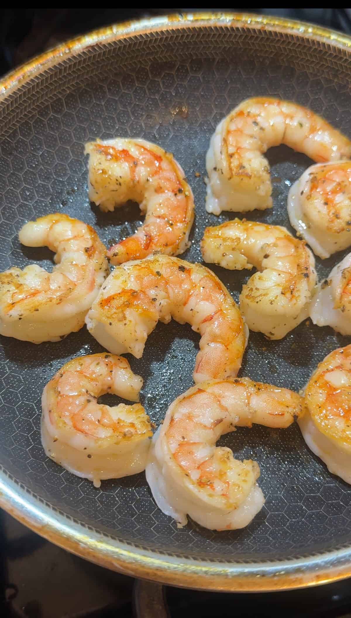 Several shrimp are being cooked in a pan, showing a pinkish-orange color with slight browning and seasoning. The shrimp are arranged in a single layer on the dark, textured surface of the pan.