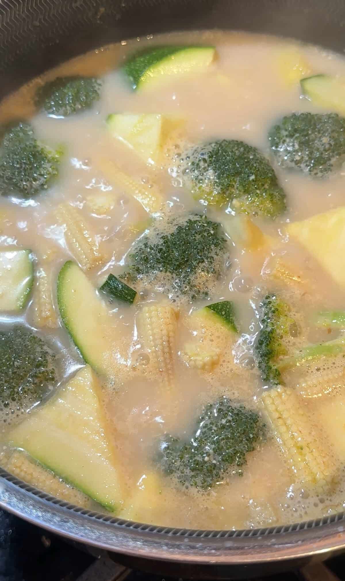 A close-up of a simmering soup with broccoli, baby corn, zucchini, and pineapple chunks in a light, creamy broth with bubbles on the surface.