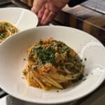 A hand holds a white bowl filled with pasta topped with a tomato-based sauce and chopped fresh herbs, resting on a counter near a wooden cutting board.