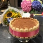 A round tiramisu cake with ladyfingers on the edge tied with a pink ribbon sits on a glass stand. In the background, there are two bouquets of colorful flowers, including sunflowers and hydrangeas.