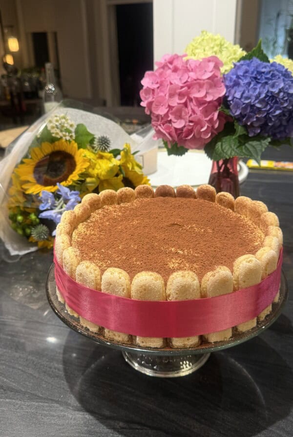 A round tiramisu cake with ladyfingers on the edge tied with a pink ribbon sits on a glass stand. In the background, there are two bouquets of colorful flowers, including sunflowers and hydrangeas.