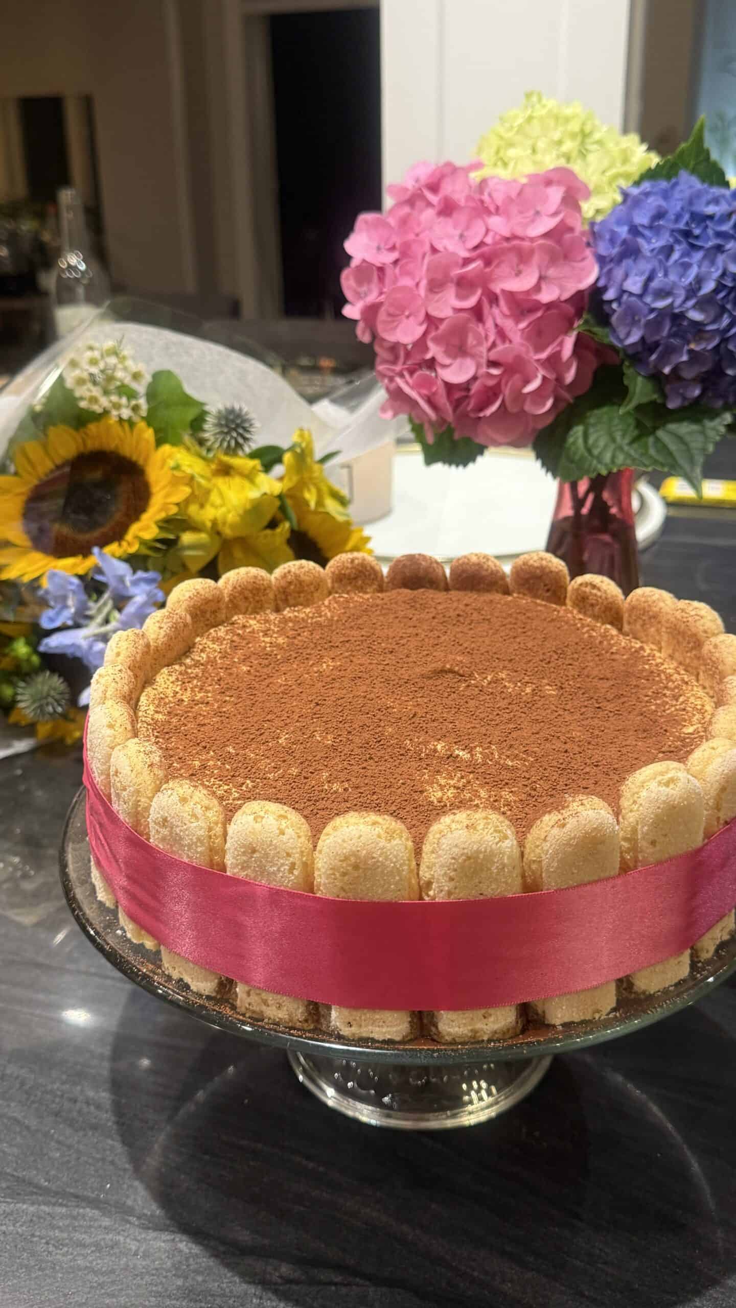 A round tiramisu cake with ladyfingers on the edge tied with a pink ribbon sits on a glass stand. In the background, there are two bouquets of colorful flowers, including sunflowers and hydrangeas.