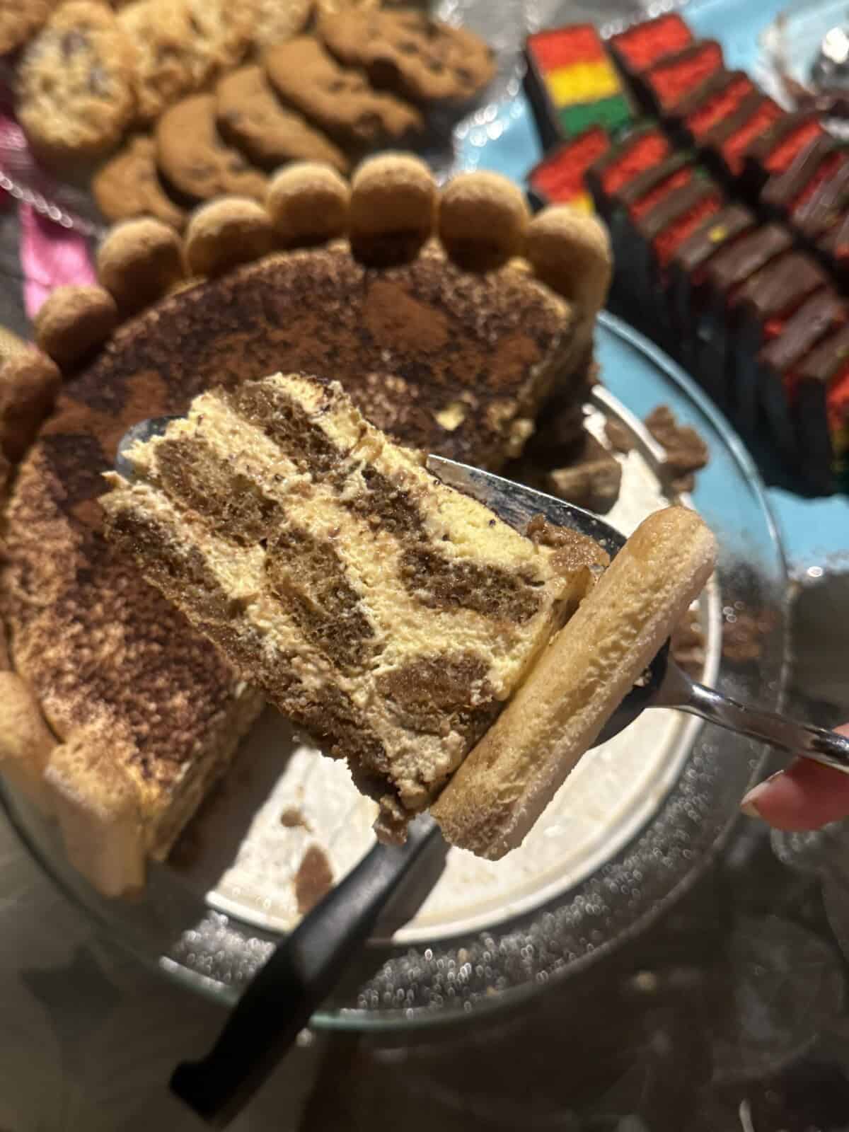A close-up of a slice of tiramisu with layers of cream and cocoa powder on a spatula held above the remaining cake, surrounded by cookies and colorful layered dessert bars on a table.