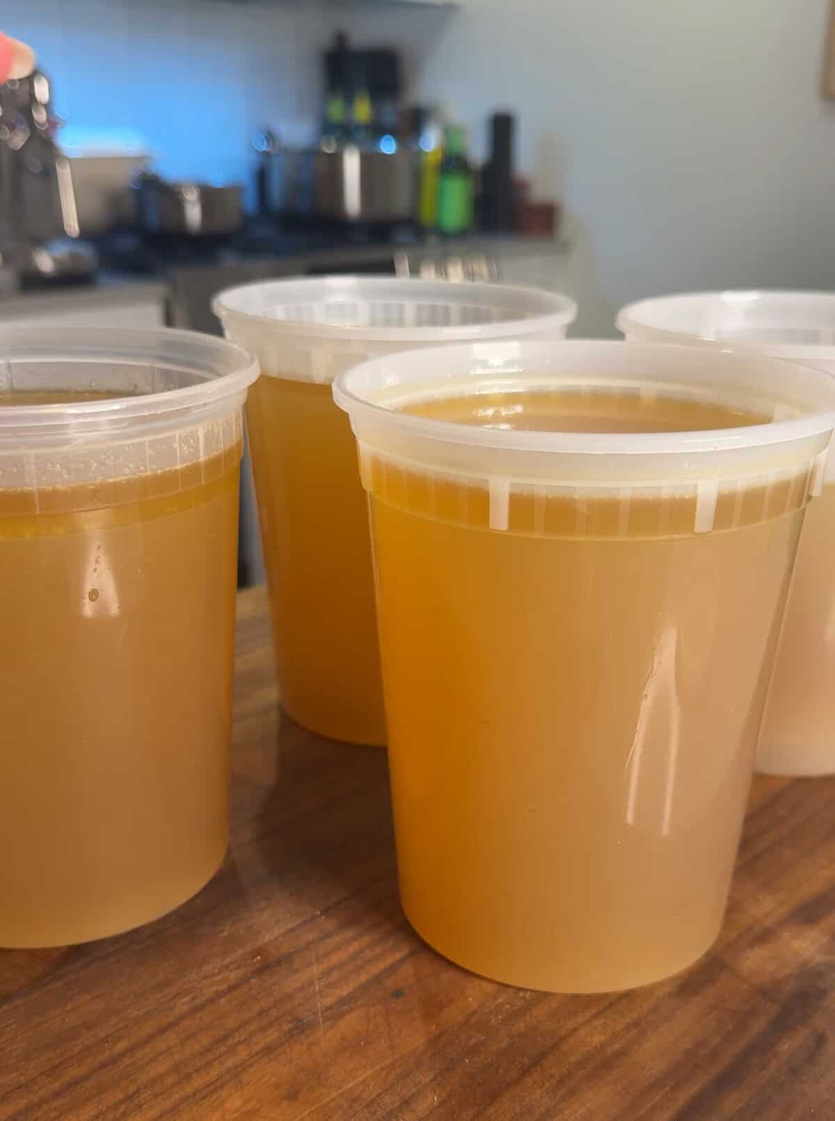 Four clear plastic containers filled with golden-brown broth are arranged on a wooden countertop in a kitchen setting with blurred pots and appliances in the background.