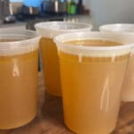 Four clear plastic containers filled with golden-brown broth are arranged on a wooden countertop in a kitchen setting with blurred pots and appliances in the background.