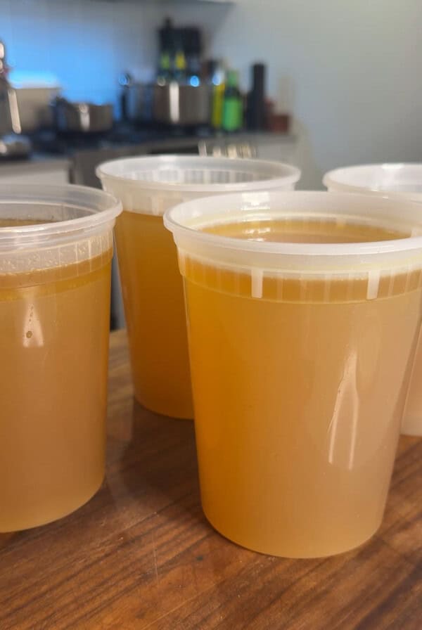 Four clear plastic containers filled with golden-brown broth are arranged on a wooden countertop in a kitchen setting with blurred pots and appliances in the background.