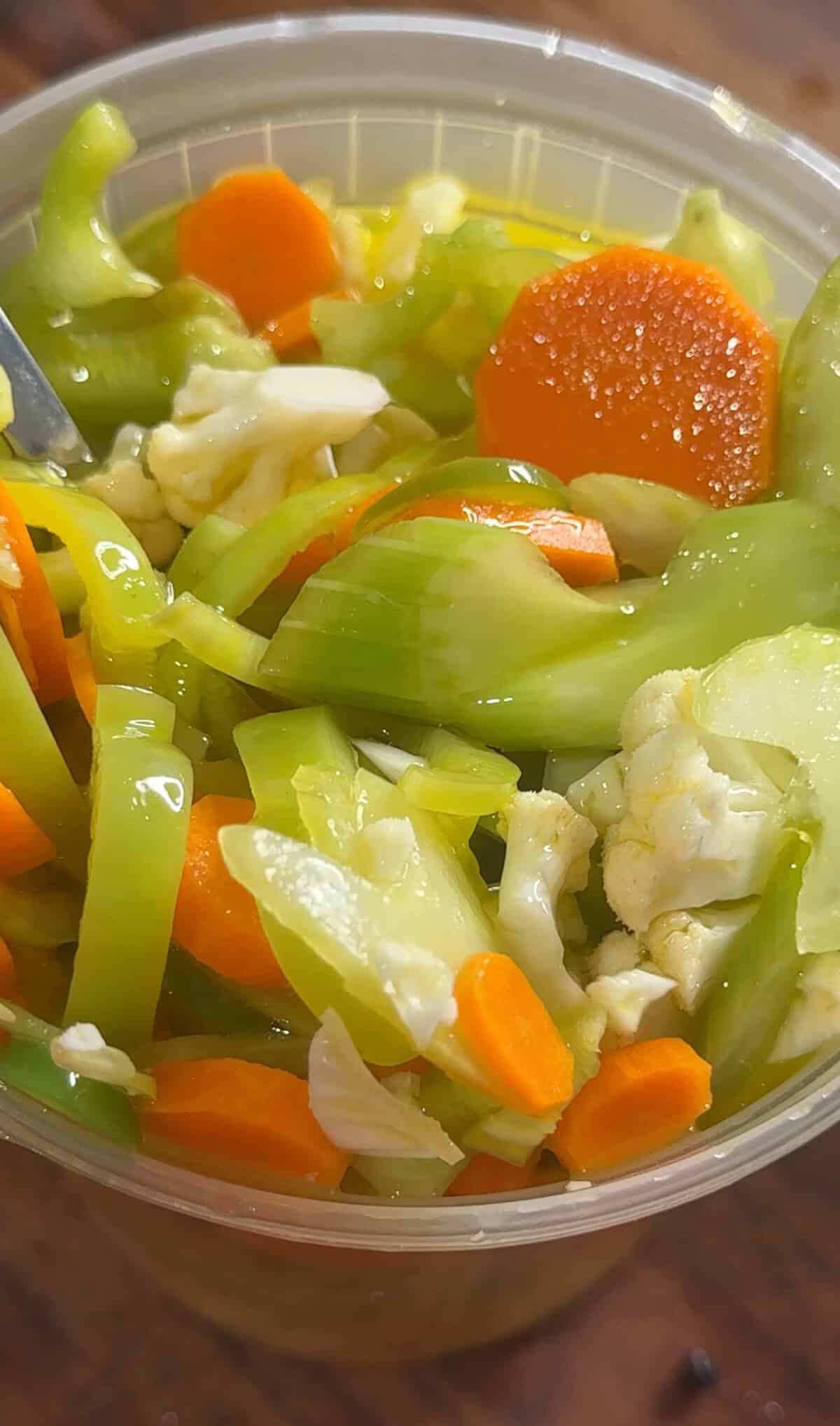 Close-up of a plastic container filled with pickled vegetables, including sliced carrots, celery, cauliflower, and green peppers, with a fork partially visible in the upper left corner.