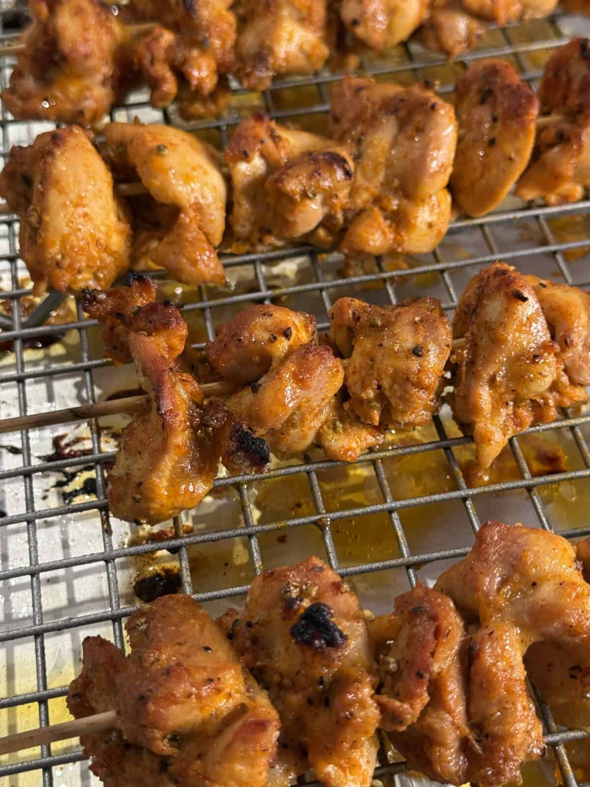 Grilled, seasoned chicken pieces on metal skewers rest on a wire rack, with golden juices below, showing a close-up of their crispy, charred texture.
