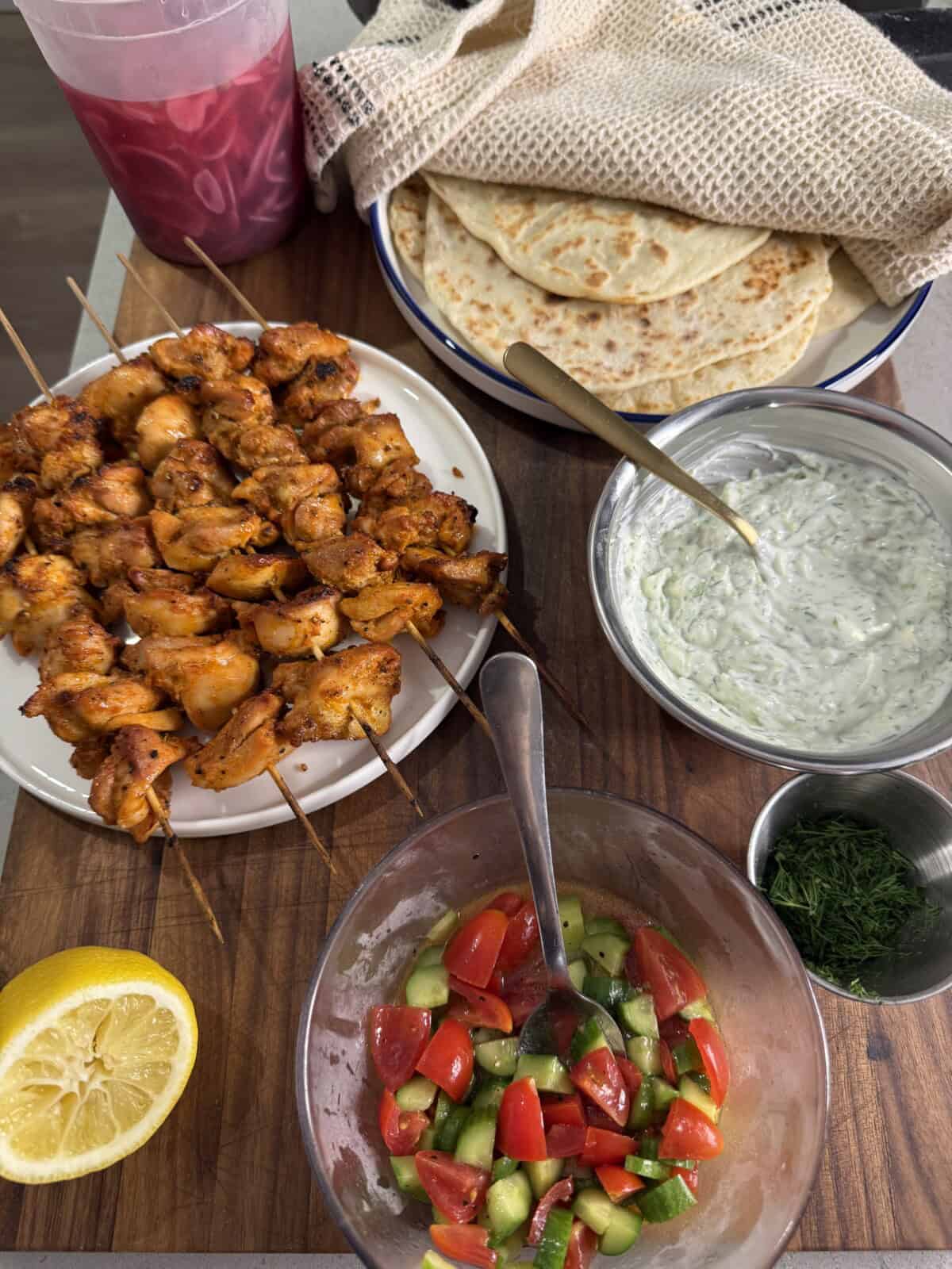 A wooden board displays grilled chicken skewers, sliced flatbreads, tzatziki sauce, chopped dill, a tomato-cucumber salad, half a lemon, and a bottle of bright red juice, with a beige cloth partially covering the bread.