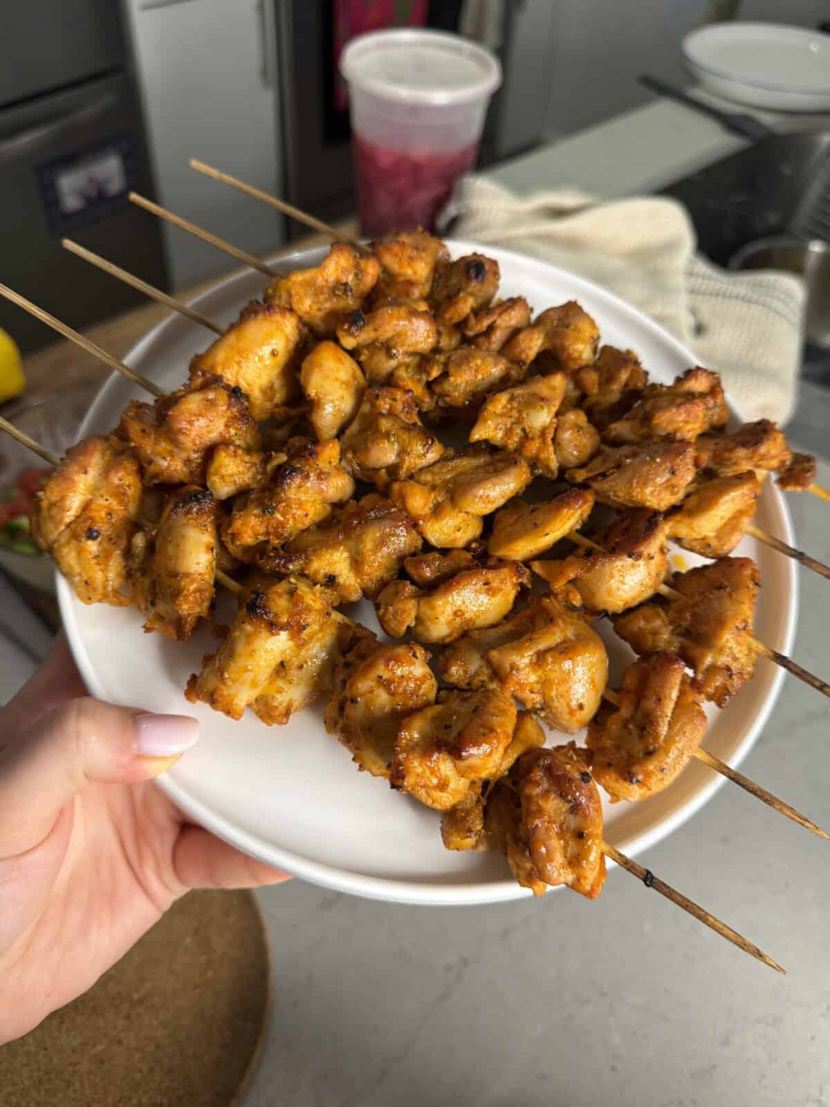 A hand holds a white plate with several skewers of grilled, seasoned chicken pieces in a kitchen setting. The chicken appears golden brown and juicy.