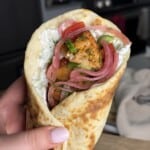 A hand holds a gyro wrap filled with grilled chicken, tzatziki sauce, pickled red onions, cucumbers, tomatoes, and fresh herbs in a soft flatbread, with a kitchen background.
