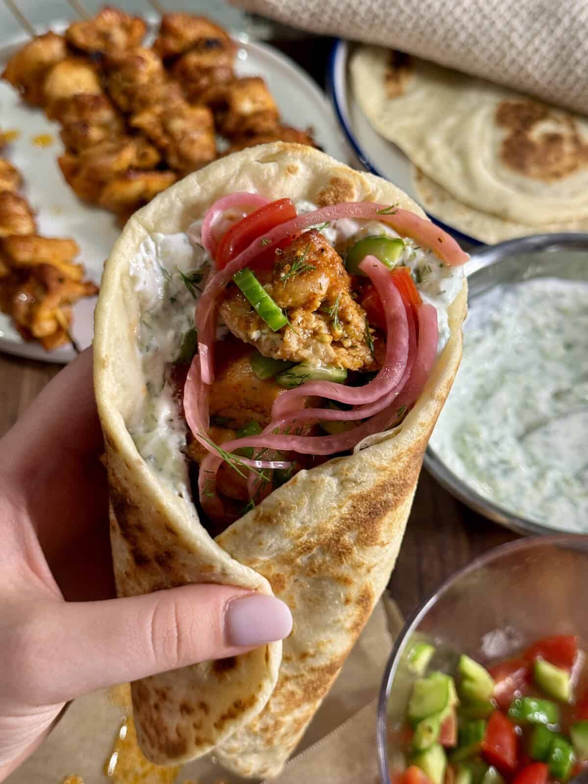 A hand holds a pita wrap filled with grilled chicken, pickled red onions, cucumber, herbs, and a creamy white sauce. Plates of chicken skewers, flatbread, sauce, and salad are visible in the background.