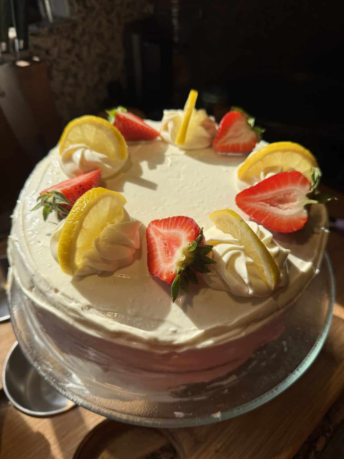 A round cake with smooth white frosting, topped with swirls of whipped cream, fresh lemon slices, and halved strawberries, displayed on a glass cake stand in natural light.