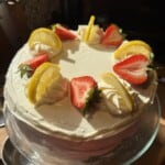 A round cake with smooth white frosting, topped with swirls of whipped cream, fresh lemon slices, and halved strawberries, displayed on a glass cake stand in natural light.