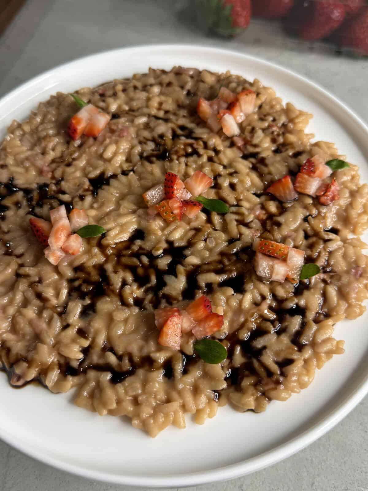 A plate of creamy risotto drizzled with balsamic glaze, topped with small pieces of fresh strawberries and a few green leaves, served on a white plate.